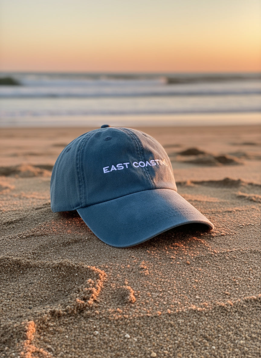 Blue cap with 'East Coast' text on a sandy beach at sunset