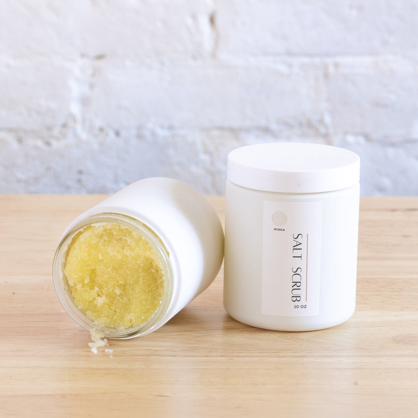 A jar of Kelp Salt Scrub is open with some scrub visible inside, placed on a wooden surface with a white brick background. The container is white with a simple design.