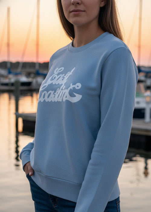 Person wearing a light blue sweatshirt with text, standing by a marina at sunset.