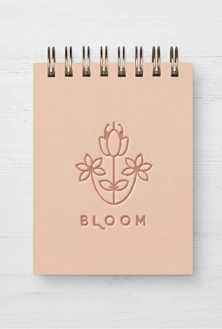 A pink mini jotter notepad with a floral design embossed on the cover and the word 'BLOOM' written below the flowers. The notepad has a spiral binding and metal bookmarks.