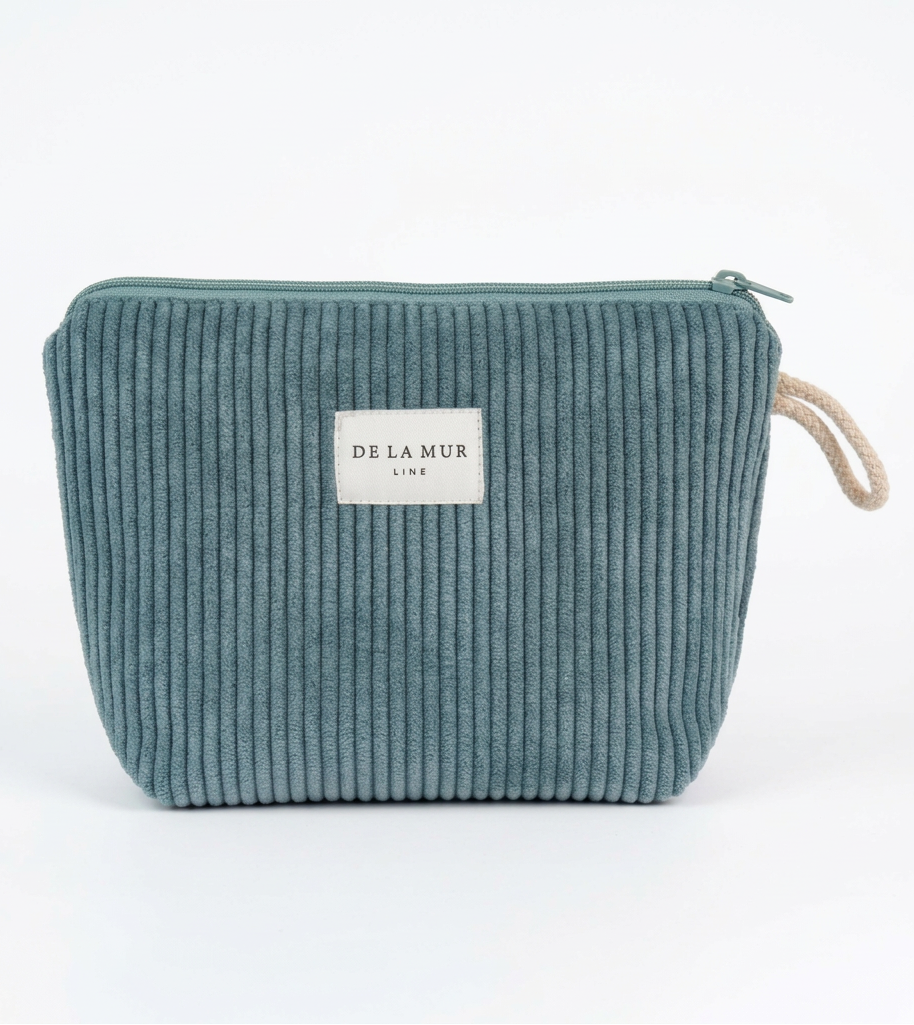 A person holds a small zip pouch in both hands. The pouch is made of a ribbed corduroy-like fabric in a muted blue-green tone and has a white rectangular label reading 'DE LA MUR LINE' on the front.