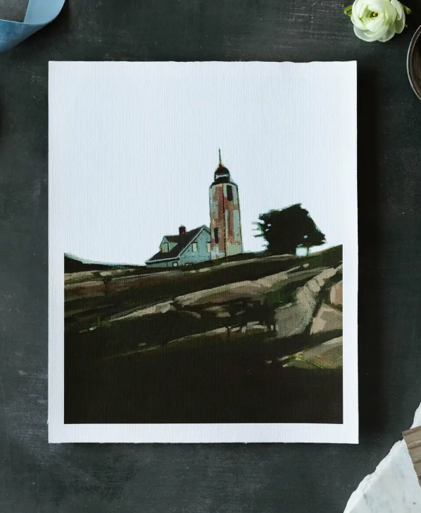 Art print of a lighthouse on a rocky shore with a white border, displayed on a dark surface.