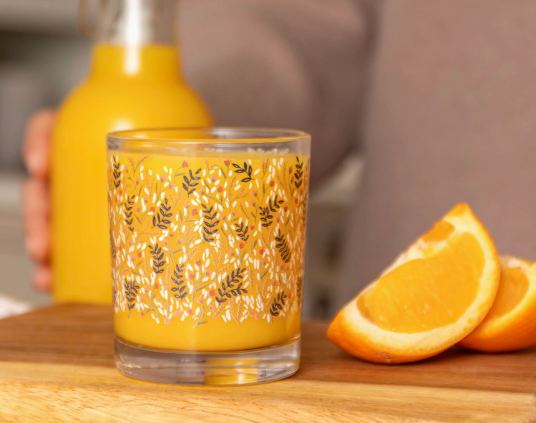 Glass with floral pattern filled with orange juice next to orange slices on a wooden surface.