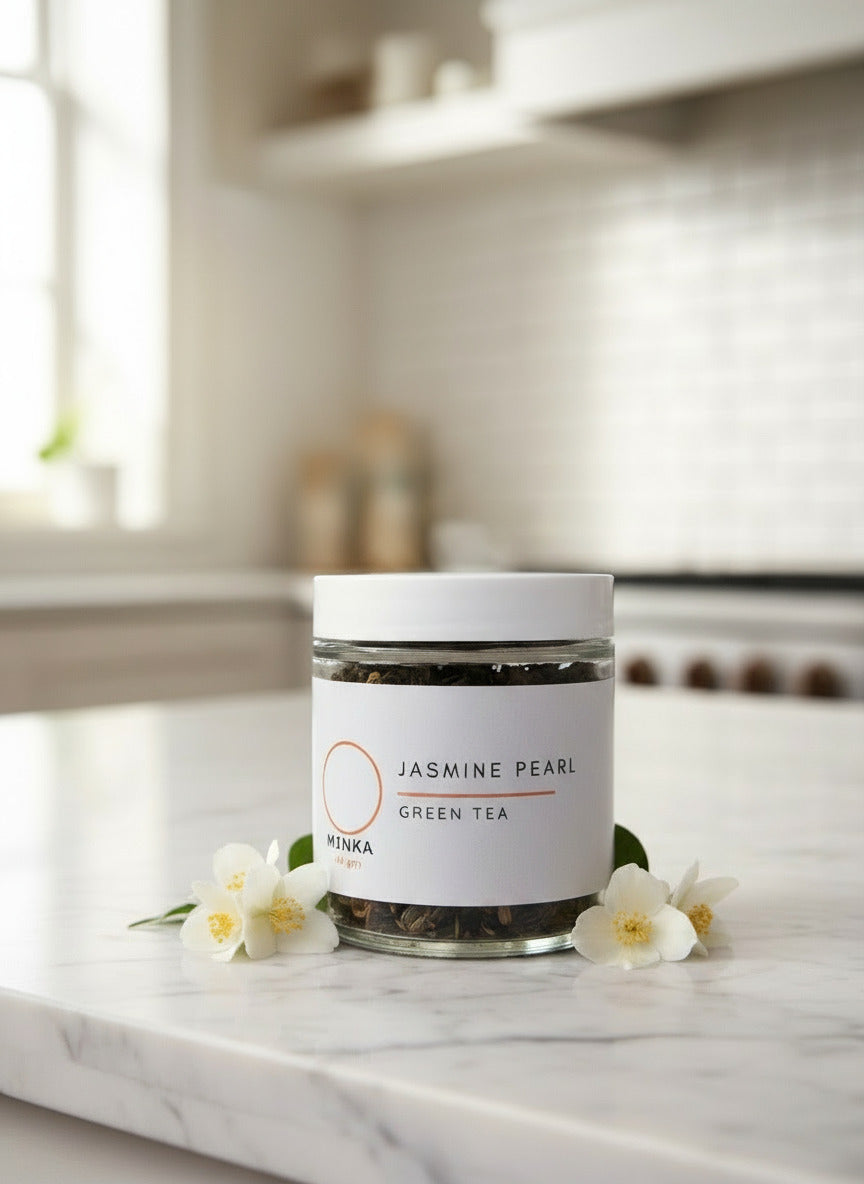 A clear glass jar containing jasmine pearl green tea with a white label that has the brand 'Minka' and the product name 'Jasmine Pearl Green Tea' written on it.