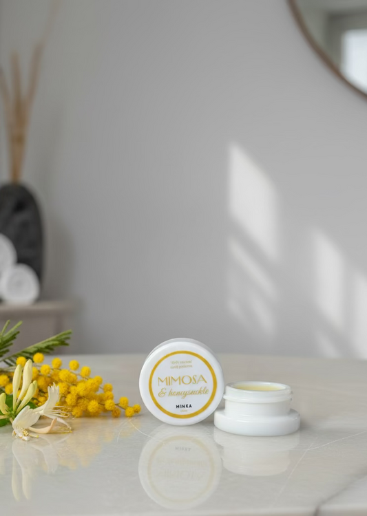 Two small containers of 'Mimosa & Honeyuckle' cream on a reflective surface with a blurred background.