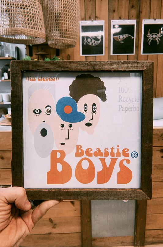 Framed poster of Beastie Boys with wooden frame held by a hand against a wooden wall background.