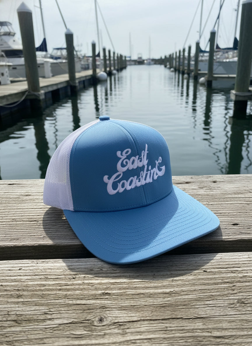 Blue and white cap with 'East Coasting' text on a wooden dock with boats in the background