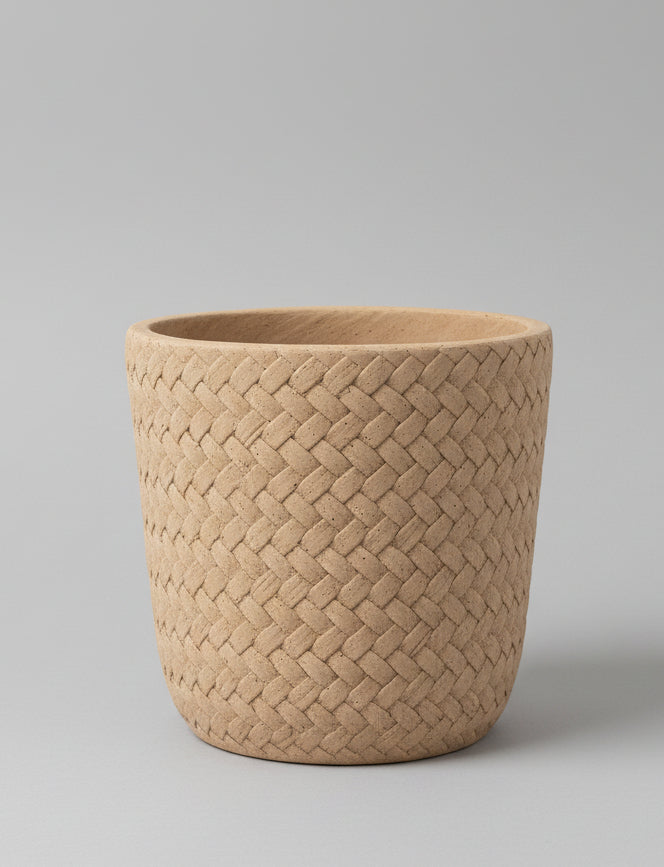 A natural woven basket planter in a light tan wood-like fiber, shown on a white surface with a wooden paneled background.