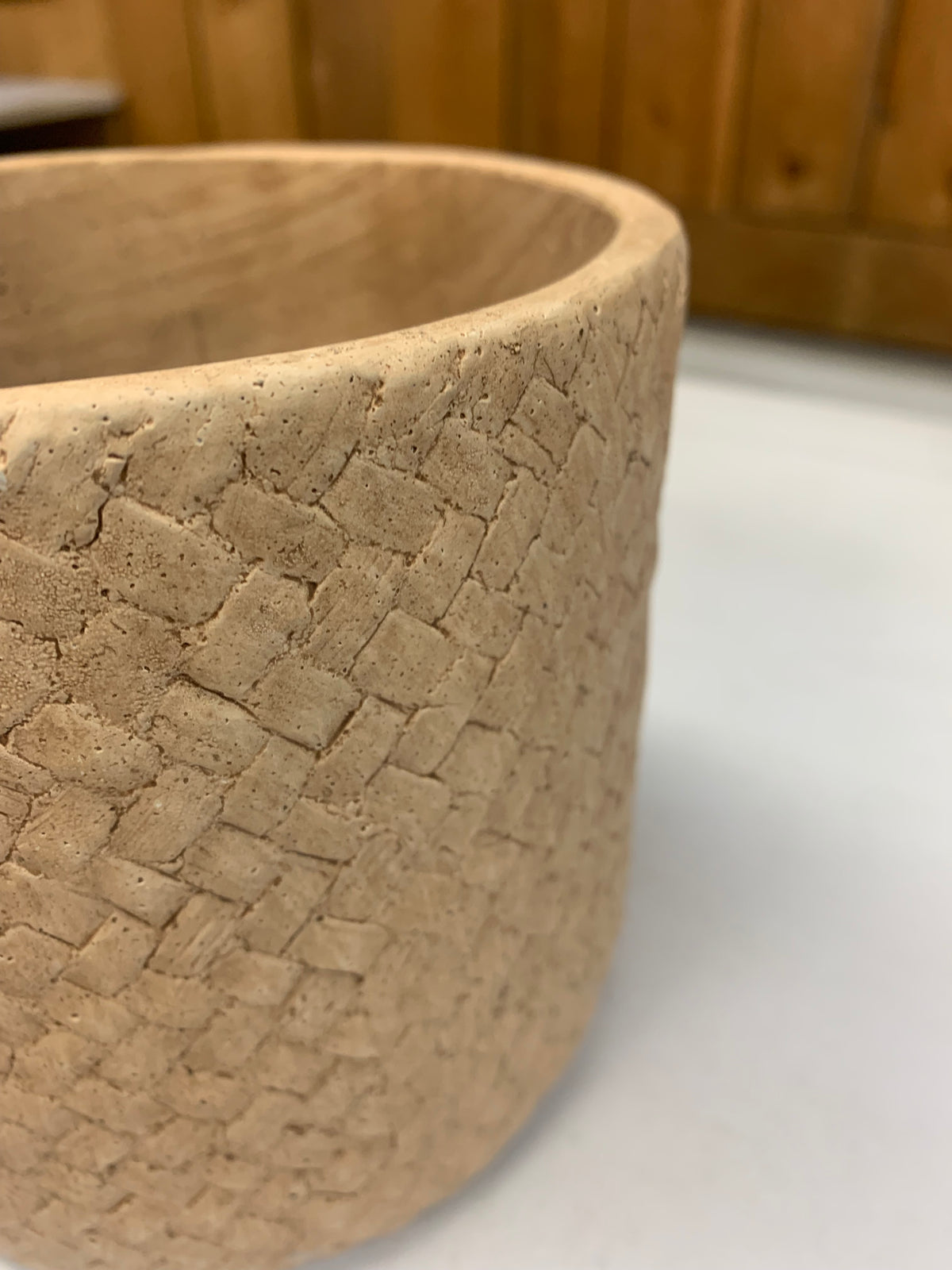 A close-up of a cylindrical planter or cup made from textured woven clay or straw material in a light tan color, set against a blurred background of a wooden wall and white surface.
