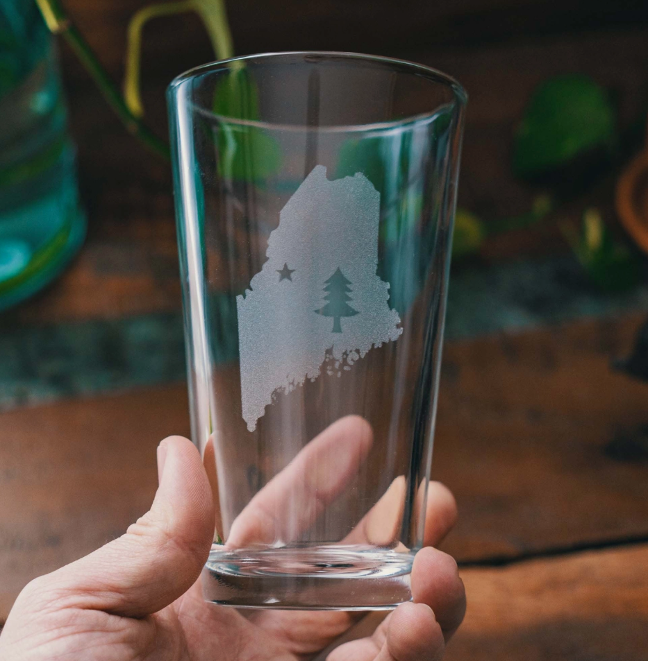 Clear glass with a engraved design of a state and tree held by a hand on a wooden surface.