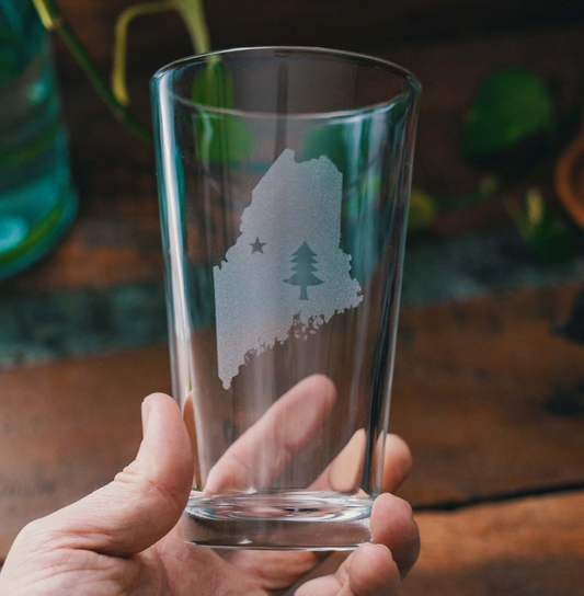 Clear glass with a engraved design of a state and tree held by a hand on a wooden surface.