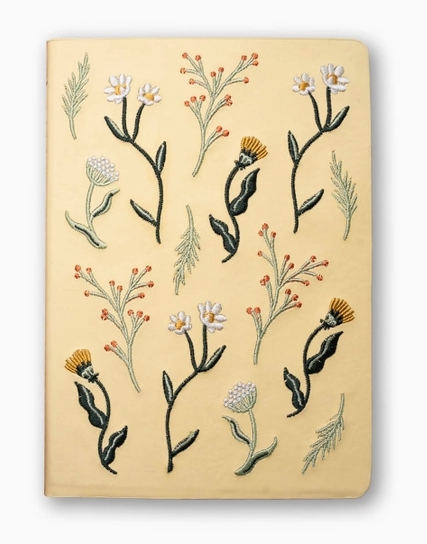 A cream-colored notebook with embroidered wildflowers and botanical stems on the cover, photographed against a clean white background.