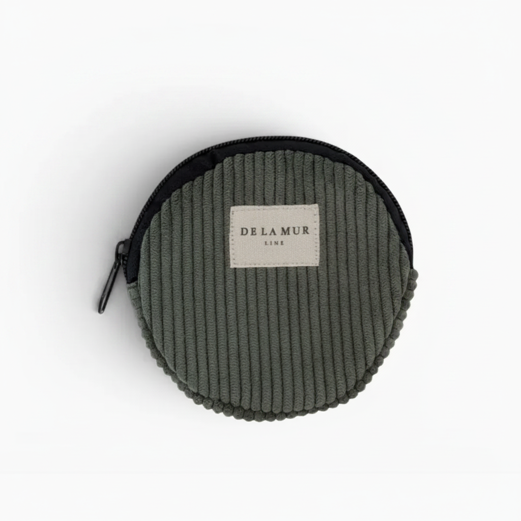 A round corduroy coin pouch in olive green with a black zipper and a beige brand label reading 'DE LA MUR LINE'.