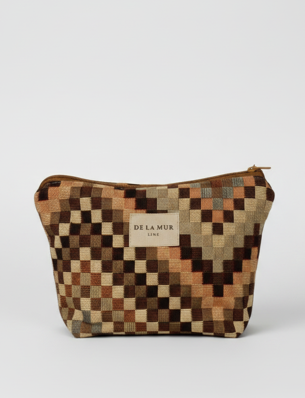 A small zippered pouch with a geometric checkered pattern in earthy tones of brown, beige, and orange. The pouch features a central beige label with the brand name 'DE LA MUR' and has a small loop handle on the right side, set against a plain white background.