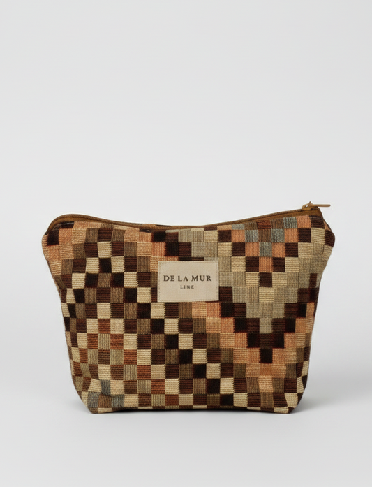 A small zippered pouch with a geometric checkered pattern in earthy tones of brown, beige, and orange. The pouch features a central beige label with the brand name 'DE LA MUR' and has a small loop handle on the right side, set against a plain white background.