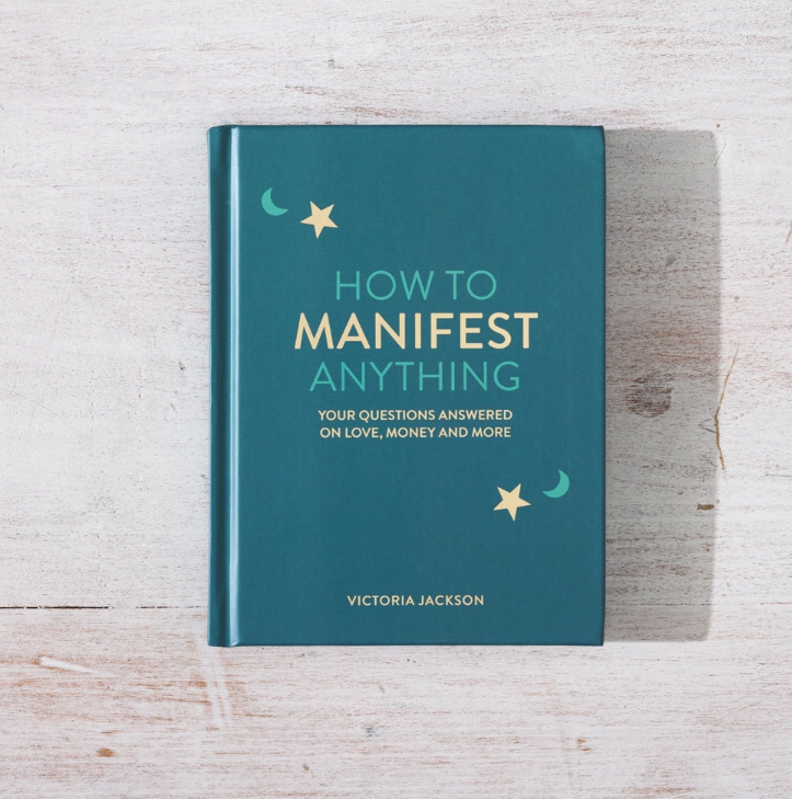 A hardcover book titled 'How to Manifest Anything' by Victoria Jackson, featuring a teal cover with gold and light blue text and celestial illustrations, placed on a light wood background.