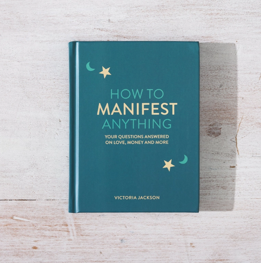 A hardcover book titled 'How to Manifest Anything' by Victoria Jackson, featuring a teal cover with gold and light blue text and celestial illustrations, placed on a light wood background.