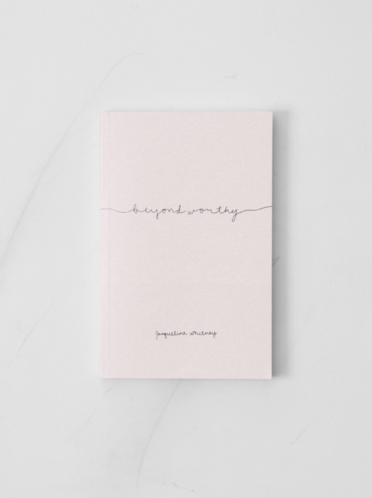 A pale pink hardcover journal with the words 'beyond worthy' in a thin gray line and 'inspiration writing' printed near the bottom, photographed on a light marble-like surface.