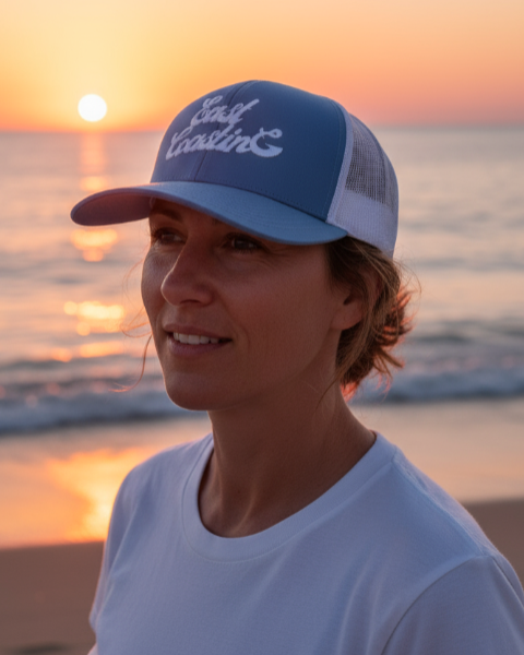 Person wearing a blue cap with 'East Coasting' text on a beach at sunset