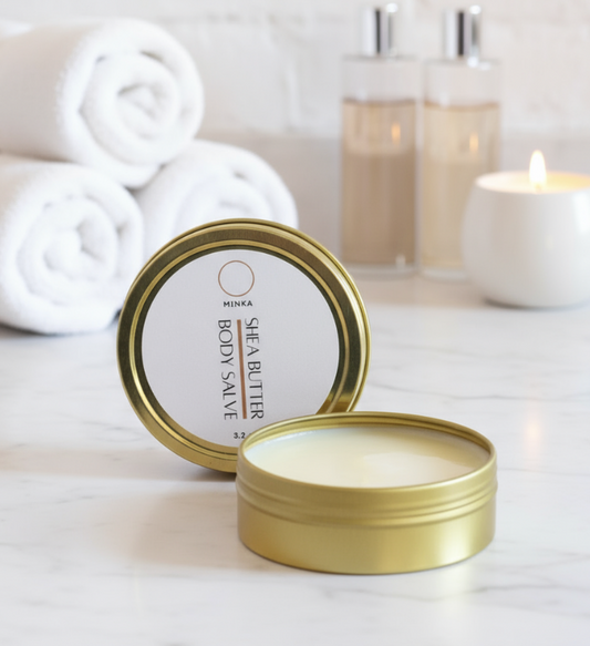 Gold tin of MINKA Shea Butter Body Salve on a white surface with towels and candles in the background