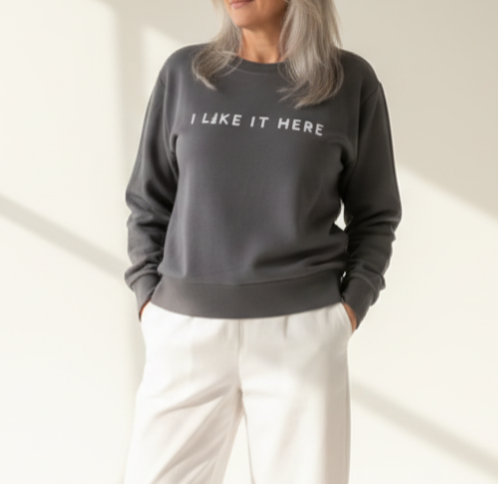 A person wearing a shadow gray sweatshirt with the text 'I like it here' printed on the front, standing outdoors with flowers in the background