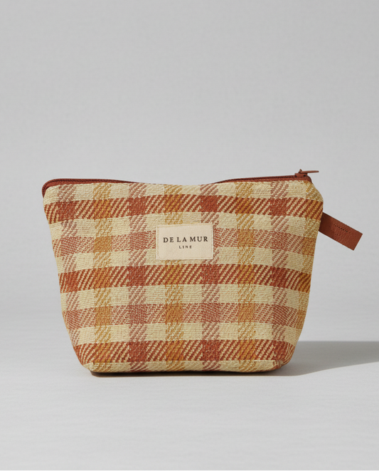 A small zippered pouch with a warm plaid pattern in rust orange, beige, and brown, featuring a central cream label that reads 'DE LA MUR LUXE'.
