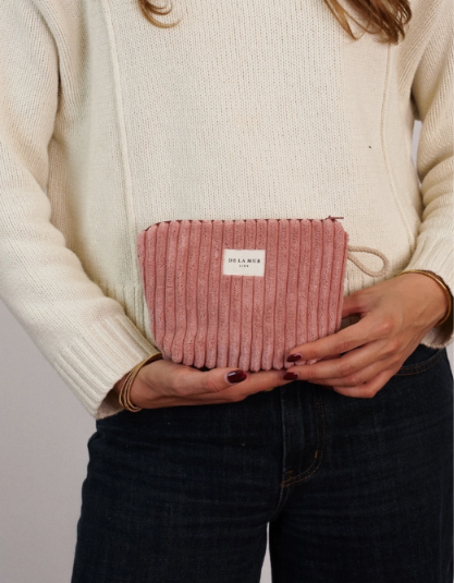 A person wearing a cream knit sweater and dark blue jeans holds a dusty pink ribbed fabric pouch with a white label that reads 'DELANEY PARIS'.