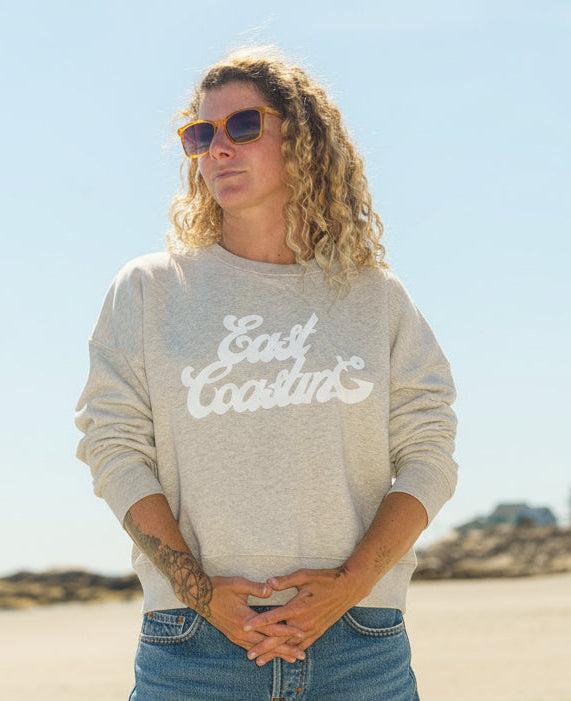 A woman standing on a sandy beach wearing a light heather gray sweatshirt with 'East Coast' printed in white script, paired with teal shorts and sunglasses.